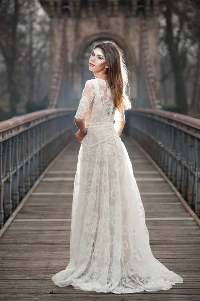 天体の光と彼女の顔に落ちる雪の梁を楽しむエレガントな白いドレスを着て素敵な若い女性 長いウェディング ドレス冬景色の橋のポーズかなりブルネットの少女 — ストック写真