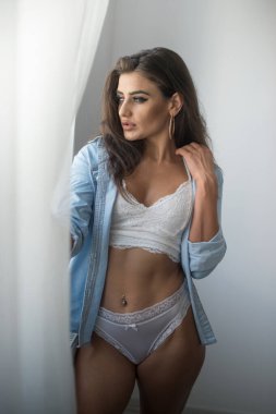 Beyaz iç çamaşırlı ve kot gömlekli çekici seksi esmer tahrik edici poz veriyor, gri bir duvarın yanında, stüdyo fotoğrafı. Klasik yatak odasında, uzun saçlı, şehvetli bir kadının portresi..