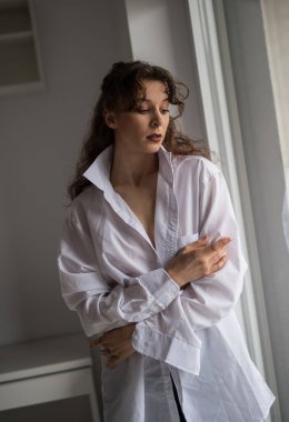 Beyaz gömlekli çekici, seksi esmer, tahrik edici poz veriyor, bir pencerenin yanında, stüdyo çekimi. Uzun saçlı, şehvetli bir kadının portresi klasik bir yatak odası sahnesinde pencereden dışarı baktı.