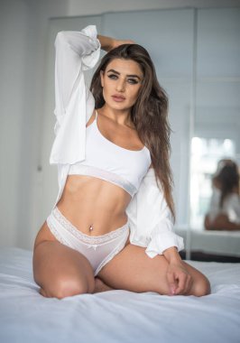 Beyaz iç çamaşırlı çekici, seksi esmer, kışkırtıcı bir şekilde poz veriyor, bir aynanın yanında, stüdyo çekimi. Yatakta beyaz sütyenli, uzun saçlı, klasik yatak odasında şehvetli bir kadının portresi..