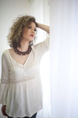Cılız, genç, kıvırcık saçlı beyaz bluz giyen bir manken. Güneş gözlüklü ve kolyeli güzel, seksi bir kadın resmi.
