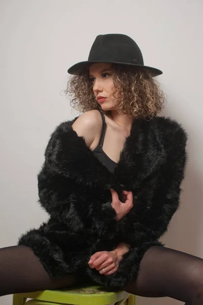 Portrait of a fashionable model with black fur sitting in a green chair in studio. young beautiful woman in black pantyhose .Portrait of attractive woman sitting in chair with black hat