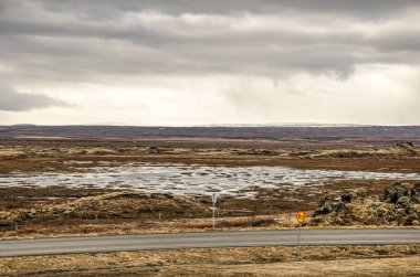 Issız bir arazide iki şerit asfalt yol sığ su birikintileri ve İzlanda 'da Myvatn Gölü' nün yanında alçak bitki örtüsü dramatik bulutlu bir gökyüzünün altında.