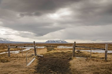 Su birikintileri ve düşük bitkilerle dolu sığ bir arazide rastgele ahşap çit ve demir kapı ve uzakta dramatik bir gökyüzünün altında Sellandafjell Dağı.