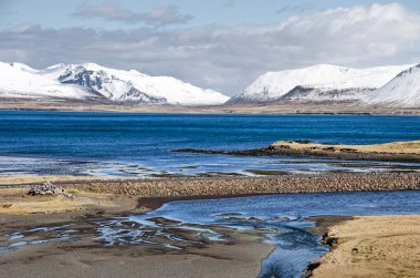 Asfalt yolu, Snaefellsnes yarımadasının kuzey kıyısındaki küçük bir nehrin kumlu deltasını geçiyor. Arka planda karla kaplı dağlar var.