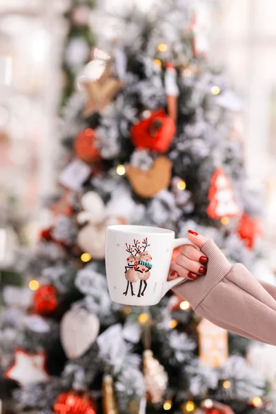 a woman's hand holds a cup against the background of a Christmas tree