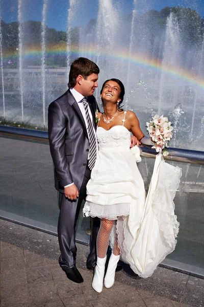 Happy bride and groom near fountain with rainbow — Stock Photo, Image