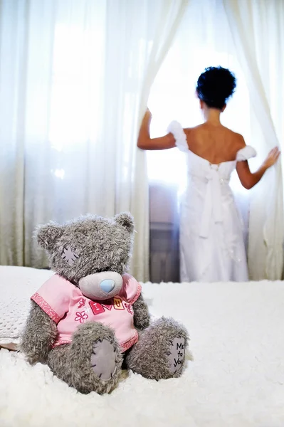 Невеста у окна и плюшевый мишка на белой кровати — стоковое фото