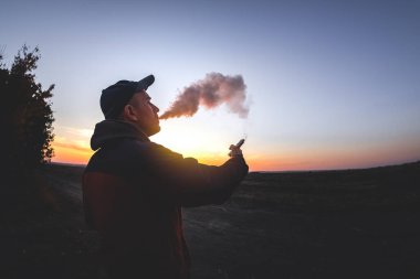 Adam günbatımında arka planda elektronik sigara içiyor.