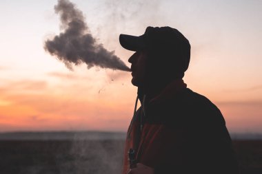 Adam günbatımında arka planda elektronik sigara içiyor.