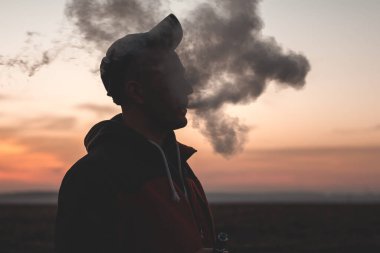 Adam günbatımında arka planda elektronik sigara içiyor.
