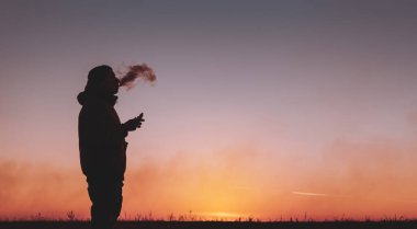 Ceketli bir adam dışarıda elektronik bir sigara içiyor. Günbatımı arka planında buhar çekmek. Analog sigaraya alternatif