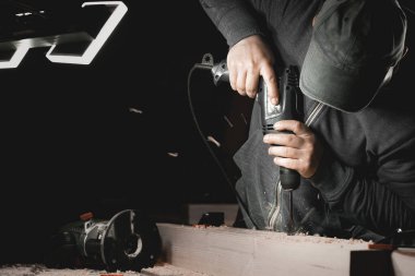 Bir adam atölyesinde matkapla çalışıyor. Marangoz, karanlık bir odada yönsel ışığı olan bir el aleti ile matkaplar.