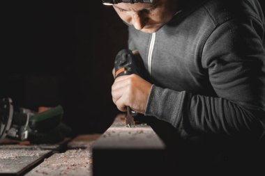 Bir adam atölyesinde matkapla çalışıyor. Marangoz, karanlık bir odada yönsel ışığı olan bir el aleti ile matkaplar.