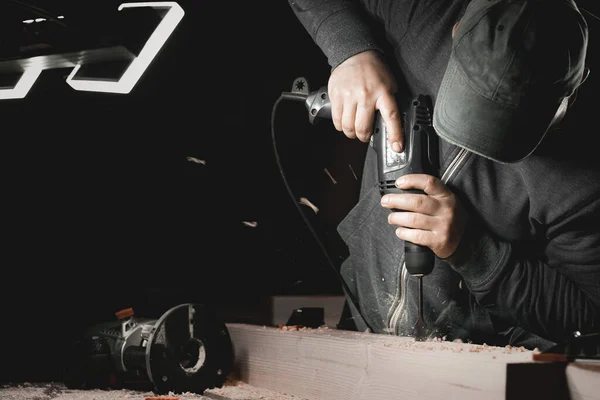 Bir adam atölyesinde matkapla çalışıyor. Marangoz, karanlık bir odada yönsel ışığı olan bir el aleti ile matkaplar.