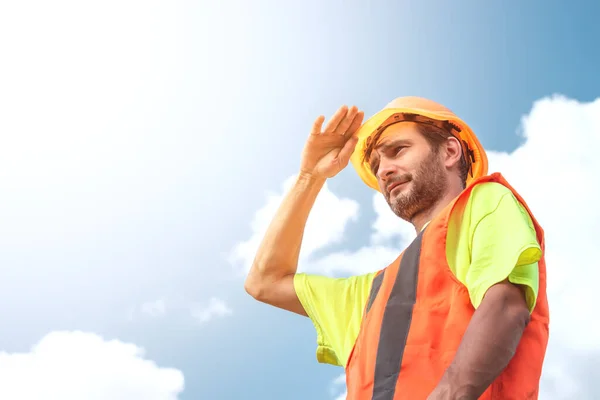 A portrait of a worker stands with confidence in an orange work suit and safety helmet against the sky. Smart industrial worker work concept.