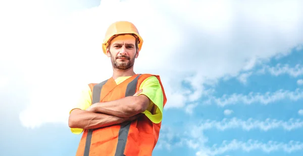 A portrait of a worker stands with confidence in an orange work suit and safety helmet against the sky. Smart industrial worker work concept.