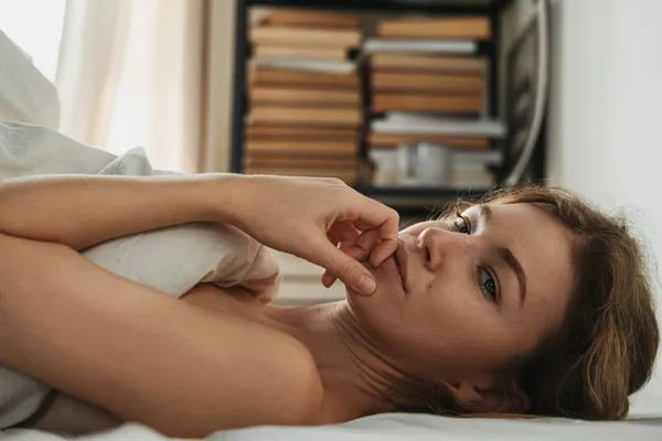 Sarı saçlı, gülümseyen genç bir kız sabahleyin yorganın altında uzanıyor ve elini ağzına tutarak kameraya bakıyor. Sağlıklı uyku konsepti.