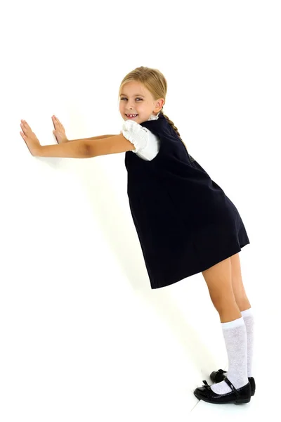 En glad blondin i en vit blus och en blå klänning lägger händerna på väggen. Härligt sjuårigt barn stående mot vit bakgrund — Stockfoto