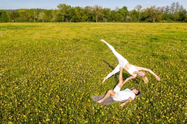 Parkta akro yoga yapan genç ve güzel bir çift. Erkek çimenlerin üzerinde yatar ve kadını ayaklarıyla dengeler..