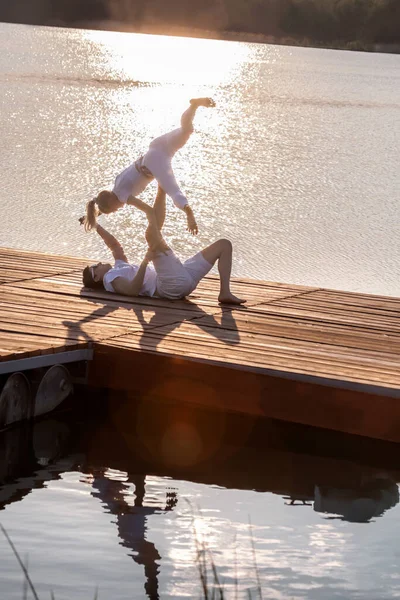 美しい若いカップルは日没に対して桟橋でアクロヨガをしています 男は桟橋に横になり 彼の足の女性のバランスをとる — ストック写真