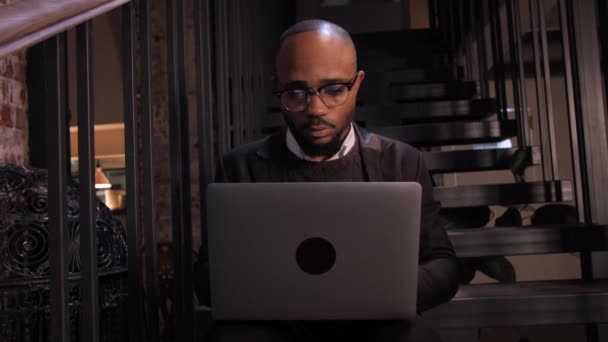 一个穿着西服，戴着眼镜的非洲年轻人坐在咖啡桌旁，坐在笔记本电脑上上网的低角度镜头 — 图库视频影像
