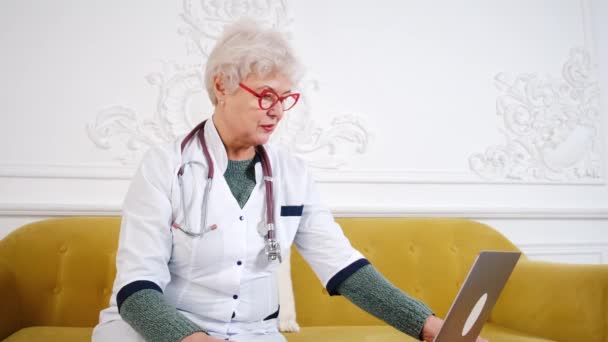 Літня жінка лікар працює за комп'ютером. Лікар телемедицини розмовляє з пацієнтом за допомогою ноутбука — стокове відео