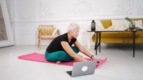 Sportig äldre kvinna som gör yoga medan du tittar på handledning på laptop — Stockvideo