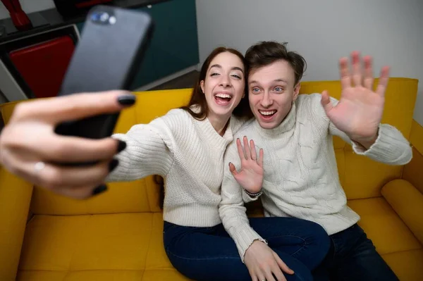 Um jovem casal em casa. Sentam-se no sofá e tiram selfies ou falam sobre comunicação por vídeo. — Fotografia de Stock