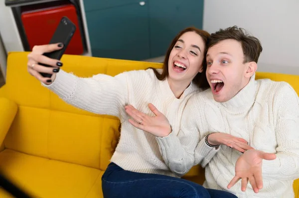 Um jovem casal em casa. Sentam-se no sofá e tiram selfies ou falam sobre comunicação por vídeo. — Fotografia de Stock