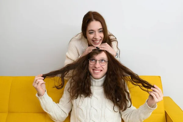 Um jovem casal passa um tempo em casa juntos. A brincar sentado no sofá. Um homem experimenta um cabelo de meninas — Fotografia de Stock