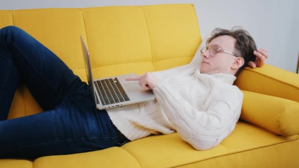 Portrait d'un bel homme avec des lunettes, à l'aide d'un ordinateur portable, assis dans le salon dans un appartement. Une personne joyeuse travaille à la maison, fait des achats sur Internet — Video