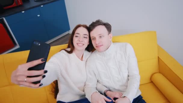 Positivo jovem casal tomando selfie enquanto sentado no sofá em casa — Vídeo de Stock