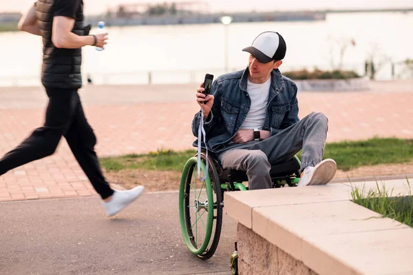 Un joven con discapacidad está sentado en un banco junto a una silla de ruedas Fotos De Stock