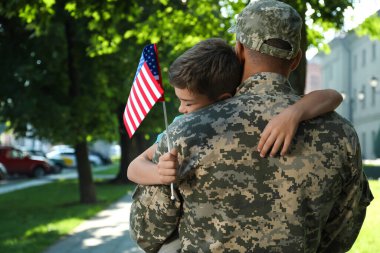 Soldier and his little son with flag of USA hugging outdoors clipart