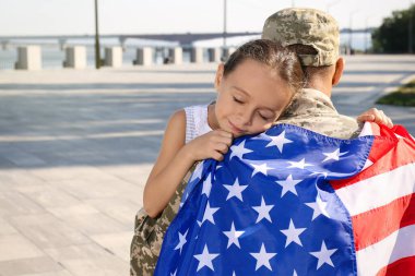 Amerikan bayrağı taşıyan asker ve küçük kızı açık havada kucaklaşıyorlar, mesaj için yer var.