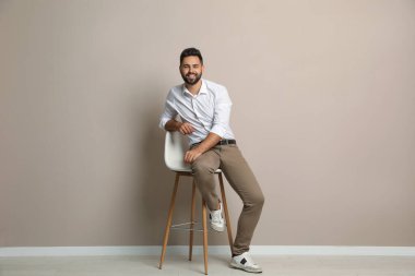 Bej duvarın yanındaki taburede oturan yakışıklı genç adam.