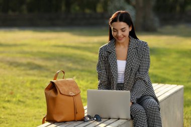 Parktaki bankta şık sırt çantalı ve dizüstü bilgisayarı olan güzel genç bir kadın.