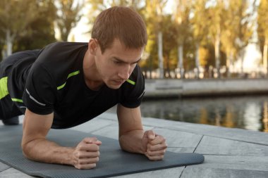 Sportif adam nehir kıyısındaki minder üzerinde kalas egzersizi yapıyor.