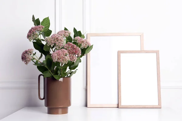 白い壁の近くのテーブルの上に美しい花と空白のフレームを持つスタイリッシュなセラミック花瓶 — ストック写真