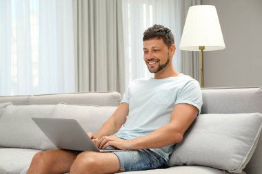 Evdeki kanepede dizüstü bilgisayar kullanan mutlu bir adam. İnternet alışverişi