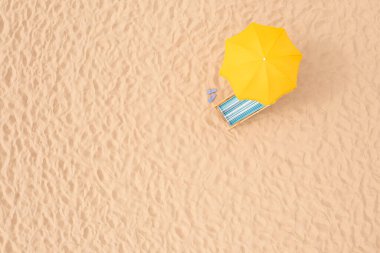 Sarı plaj şemsiyesi, kumlu sahilde güneş yatağı ve parmak arası terlikler, hava manzarası. Metin için boşluk