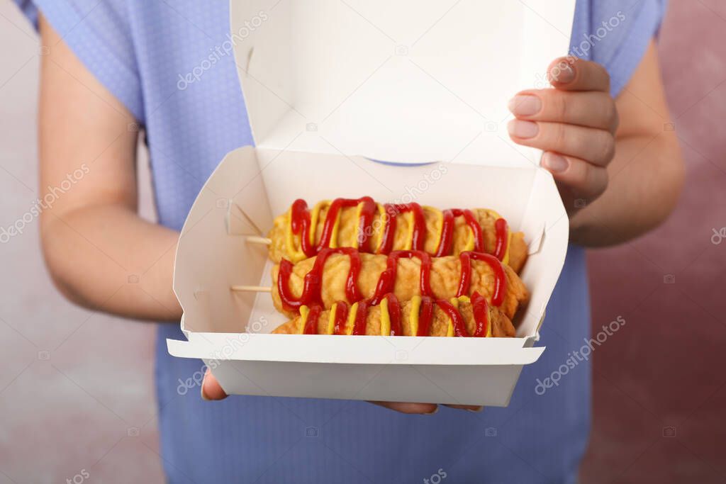 Woman holding box of delicious corn dogs with mustard and ketchup on pink background, closeup