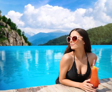 Lüks otellerde açık havuzda kokteyl içen ve güneşli bir günde güzel dağ manzaralı genç bir kadın.