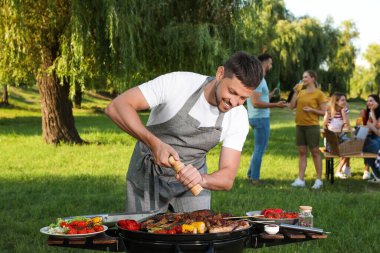 Adam parkta barbekü ızgarasında et ve sebze pişiriyor.