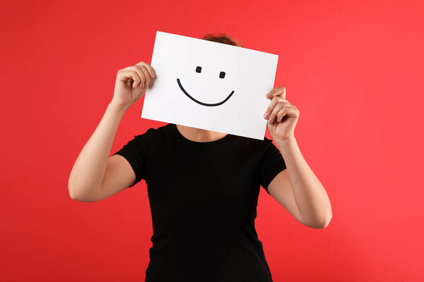 Женщина прячется за листом бумаги со счастливым лицом на красном фоне