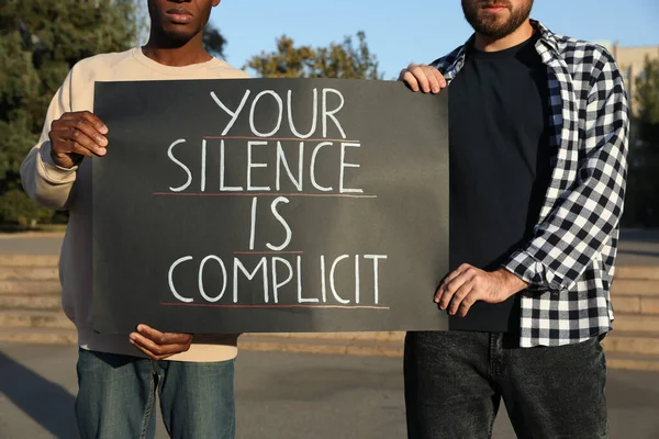 Men holding sign with phrase Your Silence Is Complicit outdoors, closeup. Racism concept