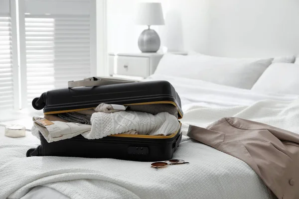 Open suitcase full of clothes, jacket and fashionable accessories on bed in room