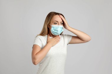 Açık gri arka planda nefes alırken acı çeken tıbbi maskeli genç bir kadın.