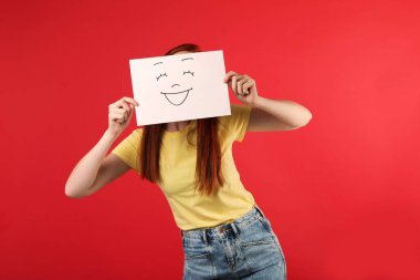 Kırmızı arka planda gülen yüzlü kâğıdın arkasına saklanan kadın.